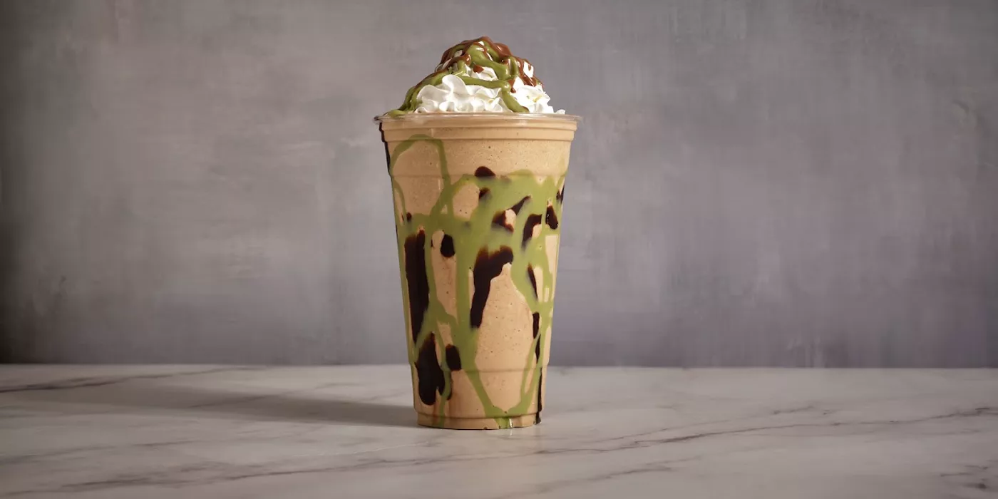 The Dubai Chocolate Frappe in a clear cup on marble: a tan blended base with pistachio and dark chocolate pulled down the inside, whipped cream and both drizzles on top.
