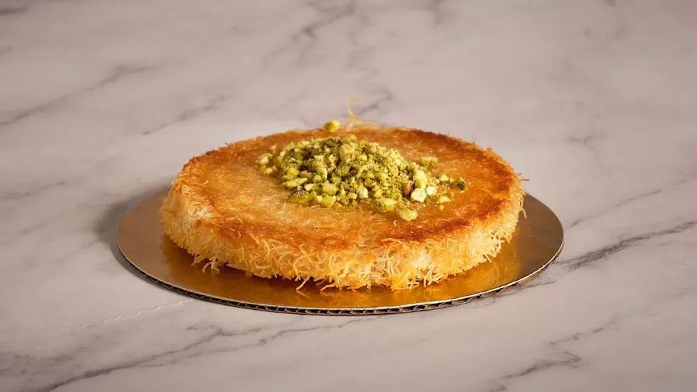 A round golden kunafa topped with a mound of crushed pistachio, on a gold board on white marble.