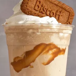 A Biscoff frappe in a clear cup, cookie butter swirled through it with whipped cream and a whole Biscoff cookie on top.