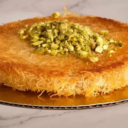 A round kunafa on a gold board, crisp golden pastry with a mound of crushed green pistachio in the middle.