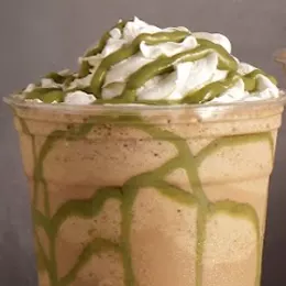 A pistachio frappe in a clear cup, whipped cream on top with green pistachio sauce drizzled over it.