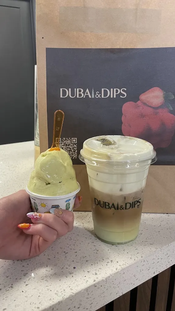 At the counter: a cup of pistachio gelato beside a layered latte in a Dubai & Dips cup, the paper takeout bag behind them.